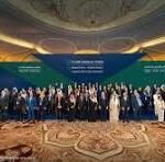 Large group photo of conference attendees in formal attire on a stage in a grand hall, with a blue banner behind them.