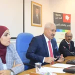 Panel of three people at a conference table; man in a dark suit with red tie speaks into a microphone, woman in a maroon hijab sits to his left, banner in Arabic behind them.