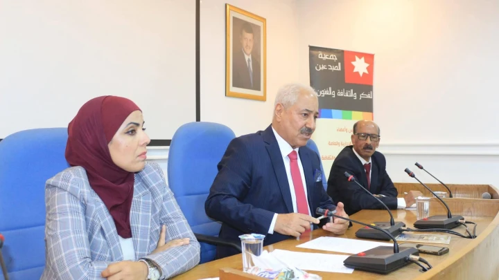 Panel of three people at a conference table; man in a dark suit with red tie speaks into a microphone, woman in a maroon hijab sits to his left, banner in Arabic behind them.