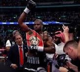 Boxer in the ring raising a championship belt in victory, with reporters and photographers around him.