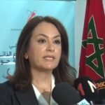 Woman in a blazer speaks into microphones at a press conference, with a Moroccan flag on the right and a blue banner behind her.