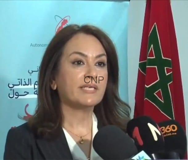 doc - Center News Network Woman in a blazer speaks into microphones at a press conference, with a Moroccan flag on the right and a blue banner behind her.
