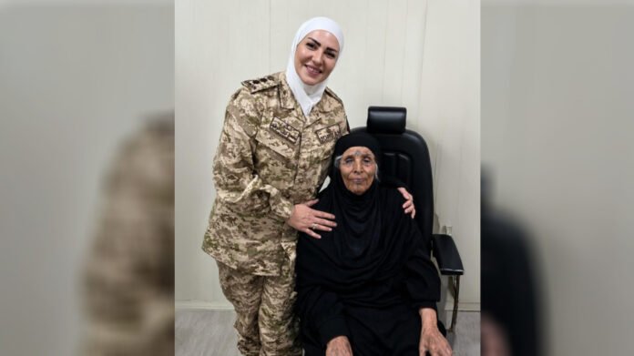 Female soldier in desert camouflage uniform and white hijab stands beside an elderly woman in black seated in a chair, both smiling indoors.