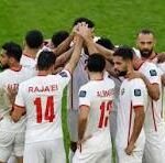 Group of football players in white jerseys celebrate in a huddle on a green pitch, raising their hands together in triumph.