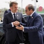 Two men in dark suits shake hands in front of a black car at a formal event, with flags in the background.