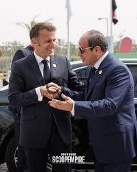 Two men in dark suits shake hands in front of a black car at a formal event, with flags in the background.