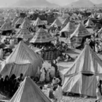 A vast desert camp with hundreds of striped canvas tents and people walking among them in the dusty terrain.