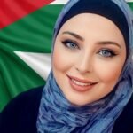 Portrait of a smiling woman wearing a blue hijab, with the Jordanian flag in the background.