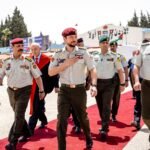 Uniformed soldiers march on a red carpet during a formal ceremony, with flags and onlookers in the background.