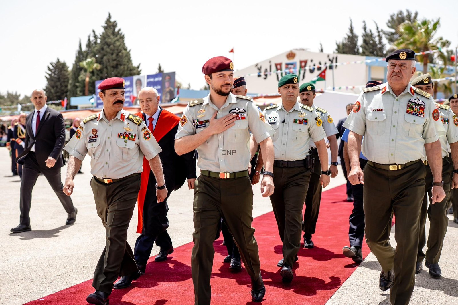 Uniformed soldiers march on a red carpet during a formal ceremony, with flags and onlookers in the background.