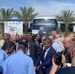 A man in a suit speaks to or greets a crowd at an outdoor event, with a bus and palm trees in the background and photographers nearby.