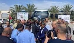 hjj - Center News Network A man in a suit speaks to or greets a crowd at an outdoor event, with a bus and palm trees in the background and photographers nearby.