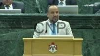 Male official delivering a speech at a lectern in a formal legislative chamber.