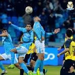 Three soccer players in blue jerseys leap to head the ball while opponents in yellow and black watch nearby, stadium background visible.