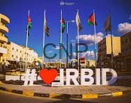 Large outdoor city square with a prominent sign featuring a heart symbol and text, flanked by several national flags and modern buildings against a bright blue sky.