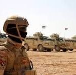 Frontline view of a soldier in desert camouflage and helmet, with a line of armored trucks in a dusty training area behind him.