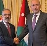 Two men in suits shake hands in a formal room with flags behind them.