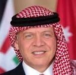 Man wearing a red-and-white checkered keffiyeh and white thobe, smiling with flags in the background.
