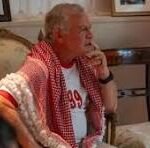Elderly man with white hair sits on a beige sofa in a living room, wearing a red shirt and a red-and-white checkered scarf, resting his chin on his hand.