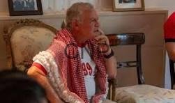 king mon - Center News Network Elderly man with white hair sits on a beige sofa in a living room, wearing a red shirt and a red-and-white checkered scarf, resting his chin on his hand.
