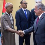 Group of men in formal and traditional attire shake hands at a diplomatic event, with a Jordan flag in the background.