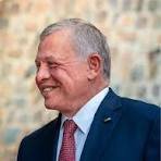 Smiling older man in a navy suit and red tie, shown in profile with a blurred stone wall background.