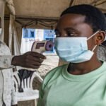 Person in a green shirt with a mask has her temperature checked with a handheld infrared thermometer under a tent, as another masked man looks on.