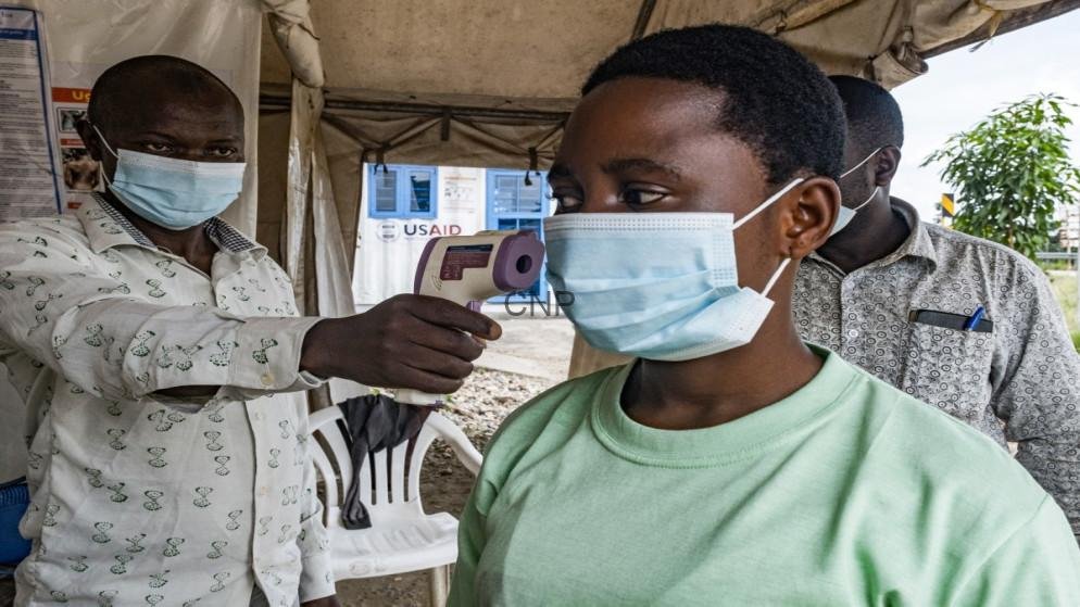 Person in a green shirt with a mask has her temperature checked with a handheld infrared thermometer under a tent, as another masked man looks on.
