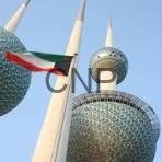 Three Kuwait Towers with a red, white, and green flag waving in the foreground against a clear blue sky.