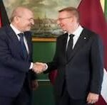 Two men in suits shake hands in front of flags during a formal meeting.