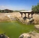 Rural scene with a small wooden pavilion over a green pond in a dry, tree-spattered landscape under a blue sky.