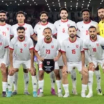 Eleven-player soccer team in white jerseys with red trim pose on the field, holding a plaque trophy; yellow-clad goalkeeper on the right, stadium crowd behind.