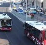 Two red city buses travel along a busy urban street with cars and pedestrians nearby.