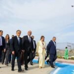 Group of formally dressed officials walking together along a paved path near a fenced facility, a Jordan flag fluttering in the background.