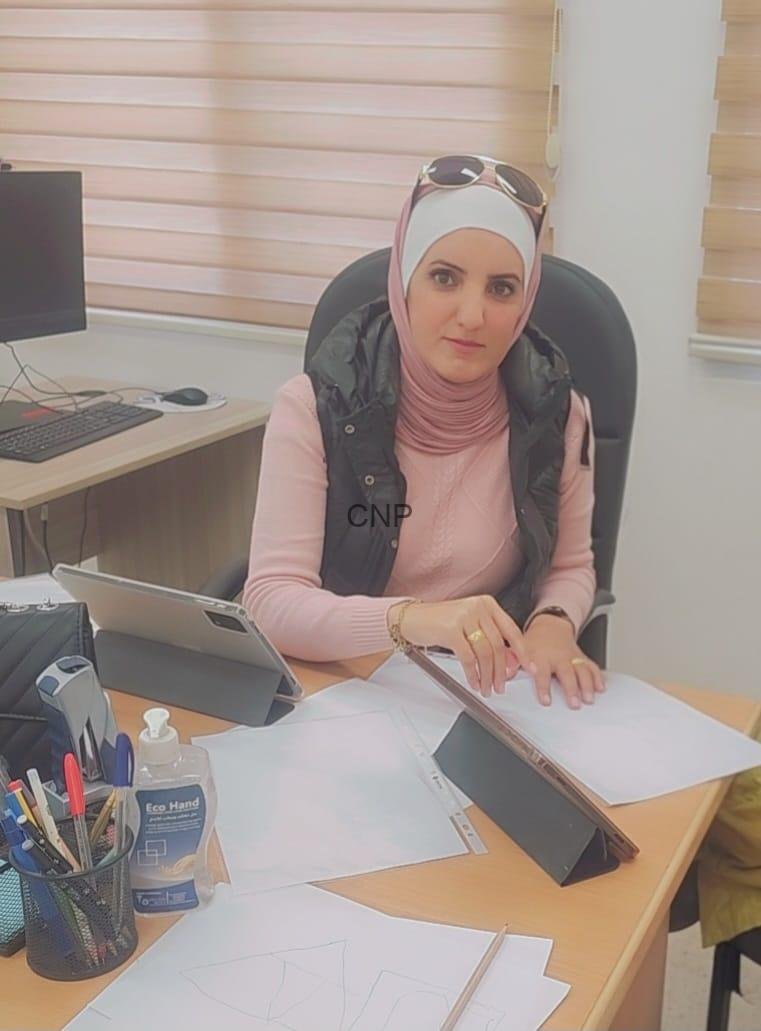 nes - Center News Network Woman wearing a pink hijab and vest sits at an office desk, reviewing papers with a tablet propped up in front of her.