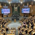 Parliament chamber with a central podium and semicircular rows of desks, flanked by large screens displaying information.
