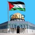 Palestinian flag flies above a historic stone building with arched entrance against a bright blue sky.