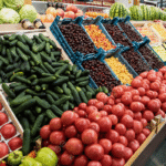 Fresh produce section with red tomatoes in the foreground, cucumbers piled beside them, and blue crates of small fruits in the middle at a supermarket.