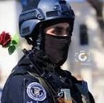 Masked police officer in tactical gear wearing a helmet and balaclava, with a red rose tucked behind his shoulder.