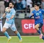 Soccer player in light blue controls the ball as a blue-clad opponent pursues.