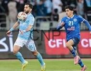 Soccer player in light blue controls the ball as a blue-clad opponent pursues.