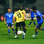 Soccer match: a yellow jersey player controls the ball while blue-jersey opponents converge on a green field under stadium lights.