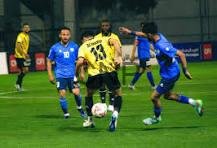 Soccer match: a yellow jersey player controls the ball while blue-jersey opponents converge on a green field under stadium lights.