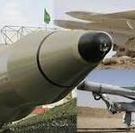 Two missiles on display outdoors: a large gray missile in the foreground and a smaller, flag-marked missile with a red tip in the background.