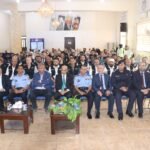 Large hall filled with people seated for a formal event; front row includes police officers in light blue uniforms.