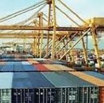 Port with stacked shipping containers and a large gantry crane over a cargo ship in the background at a busy harbor.