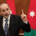 A man in a suit speaks at a podium, raising his index finger, with the Jordanian flag in the background.
