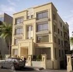 Three-story beige apartment building with balconies on a sunny street, palm trees in front and parked cars nearby.