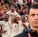 Serious coach in the foreground as a men's soccer team celebrates in white jerseys with red numbers in the background, flags visible in the stands.