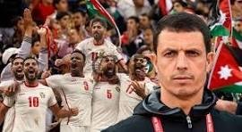 Serious coach in the foreground as a men's soccer team celebrates in white jerseys with red numbers in the background, flags visible in the stands.